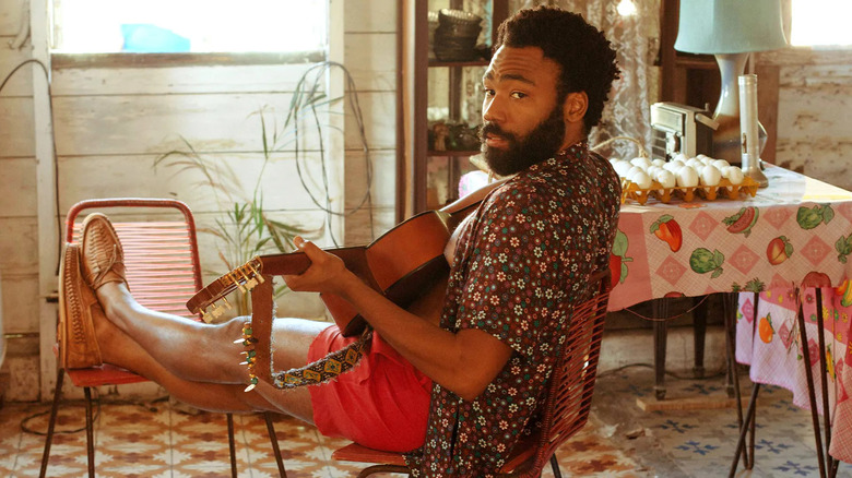 Deni Maroon sitting at his kitchen table with a guitar in his lap in Guava Island