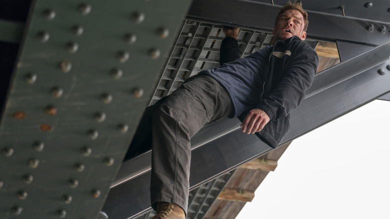 Alan Ritchson as Jack Reacher hanging from a steel girder underneath a bridge in Reacher