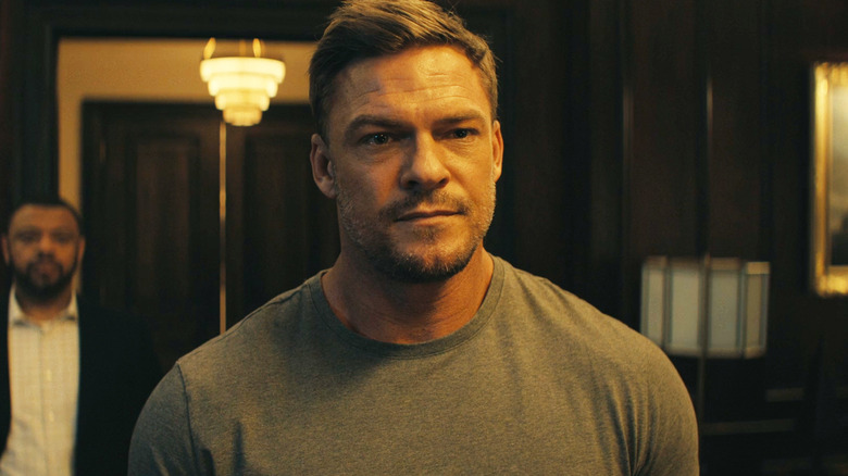 Alan Ritchson's Jack Reacher stands in an office with a bodyguard behind him in Reacher