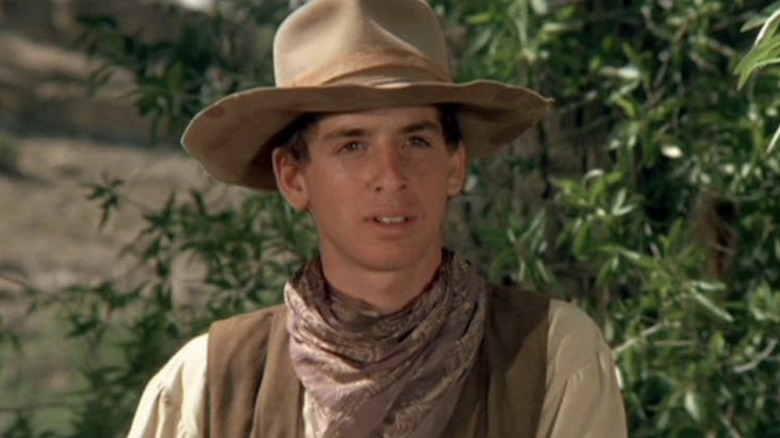 రాబర్ట్ కరాడిన్ యొక్క స్లిమ్ హనీకట్ ది కౌబాయ్స్‌లో బయట నిలబడి ఫ్రేమ్ అంతటా చూస్తున్నాడు