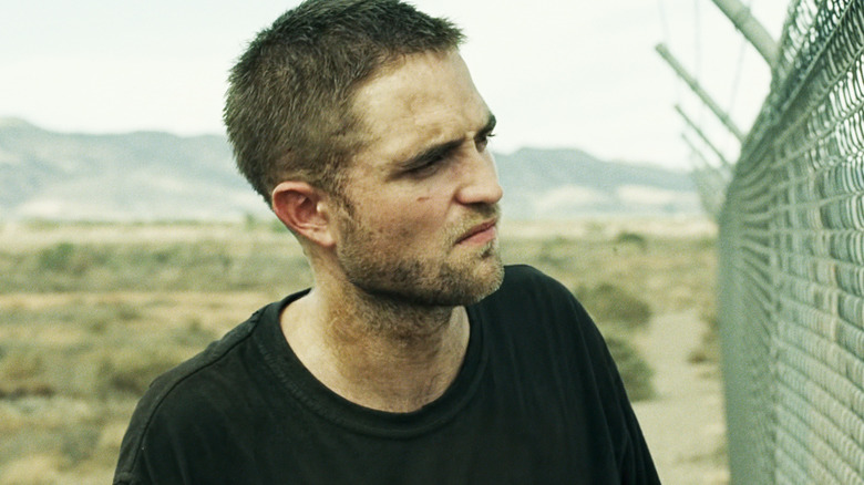 Robert Pattinson's Reynolds stands in front of a chain link fence in The Rover