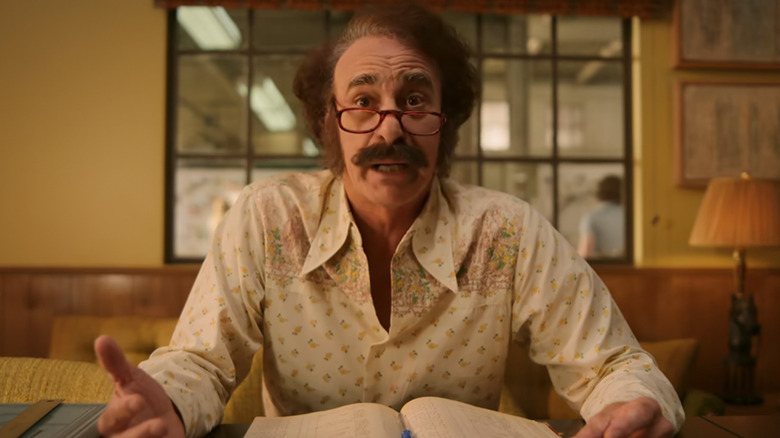 Ed Alonzo as Gerry talking at a desk with papers in Minx