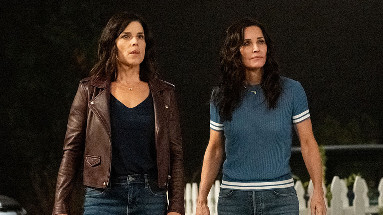 two brunette women standing next to one another, one in a brown leather jacket, black shirt and jeans and another in jeans and a blue shirt with white stripes around the arms and waist
