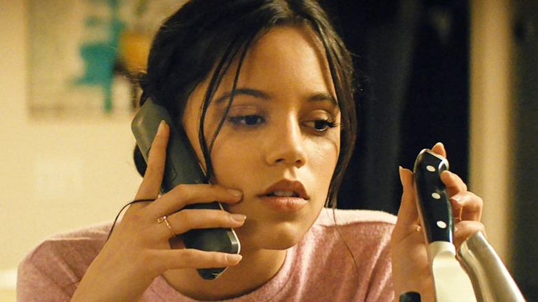 brunette woman in a pink shirt holding the handle of a knife in her left hand a phone to her ear in her right hand