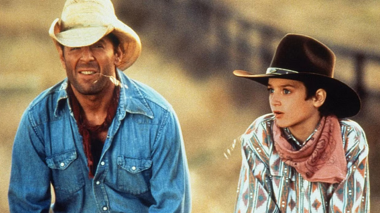 North (Elijah Wood) and the Narrator (Bruce Willis) sit outdoors wearing cowboy hats and Western outfits, looking concerned, with a dry field and wooden fence in the background