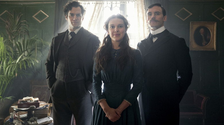 Henry Cavill as Sherlock Holmes and Sam Claflin as Mycroft Holmes standing behind Millie Bobby Brown as Enola Holmes in a poshly-decorated room in Enola Holmes
