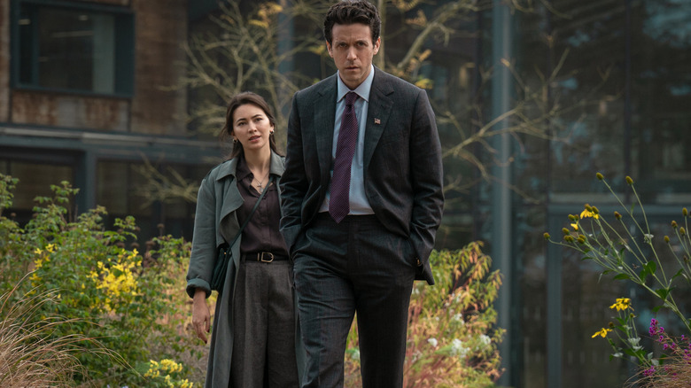 Ashley Zukerman as Congressman Daniel Keene wearing a suit and walking outside a building in front of Jessica Henwick as Helen Drew in Silo