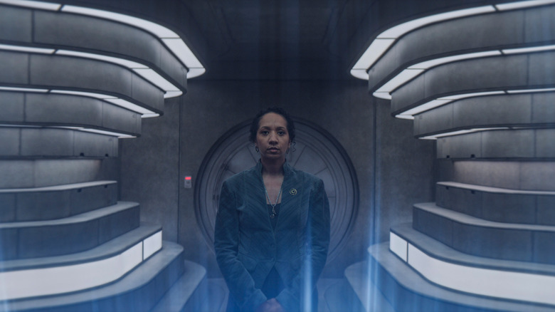 Alexandria Riley as Camille Sims standing in a brightly-lit room opposite a display of blue lights in Silo