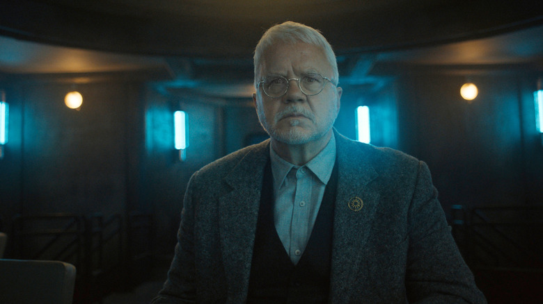 Tim Robbins as Bernard Holland wearing a suit jacket and standing in a darkly-lit room in Silo