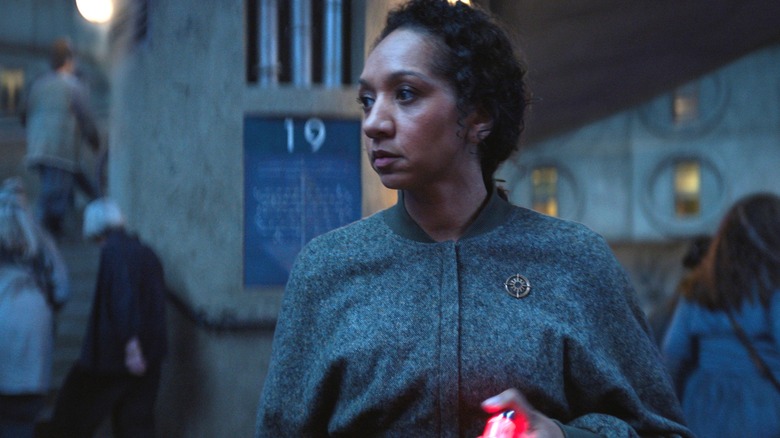 Alexandria Riley as Camille Sims standing on a crowded staircase and holding a keychain with a glowing red light in Silo