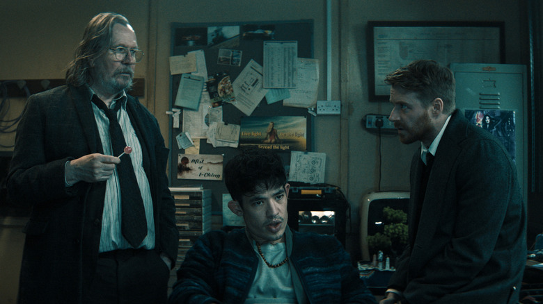 Gary Oldman as Jackson Lamb holding a lollypop and standing in a cramped room next to Christopher Chung as Roddy Ho and Jack Lowden as River Cartwright in Slow Horses