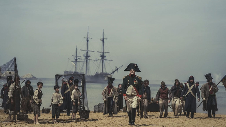 Joaquin Phoenix's Napoleon walking on a beach with lots of people and a ship in the background