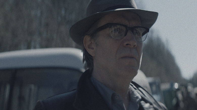 Rhys Ifans as the Chief Designer wearing a top hat and standing outside next to a line of military vehicles in Star City