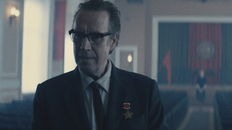 Rhys Ifans as the Chief Designer attending a ceremony and wearing a medal on his suit jacket in Star City