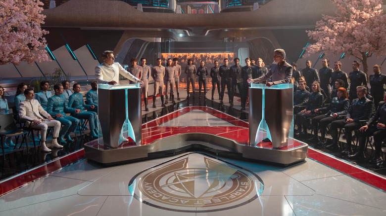 Two Starfleet commanders stand opposite each other at podiums, one dressed in white and the other in grey, while an audience watches from the sidelines in "Star Trek: Starfleet Academy"