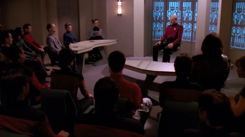 Kapitein Picard zit midden in een proefkamer, omringd door Starfleet-officieren in Star Trek: The Next Generation