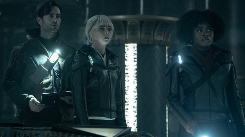 Cillian O'Sullivan as Dr. Roger Korby, Jess Bush as Chapel, and Celia Rose Gooding as Uhura standing in a dark room with concerned looks on their faces in Star Trek: Strange New Worlds