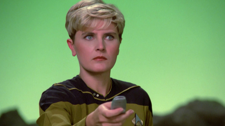 Denise Crosby as Tasha Yar holding up a handheld device and standing in front of a green background in Star Trek: The Next Generation
