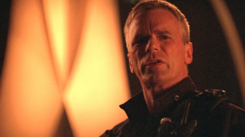 Richard Dean Anderson's Jack O'Neill looks concerned as he stands illuminated by an orange glow in Stargate SG-1