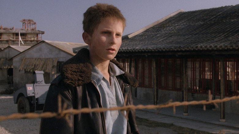 Christian Bale dans le rôle de Jamie portant une veste en cuir derrière une clôture en fil de fer barbelé dans Empire of the Sun