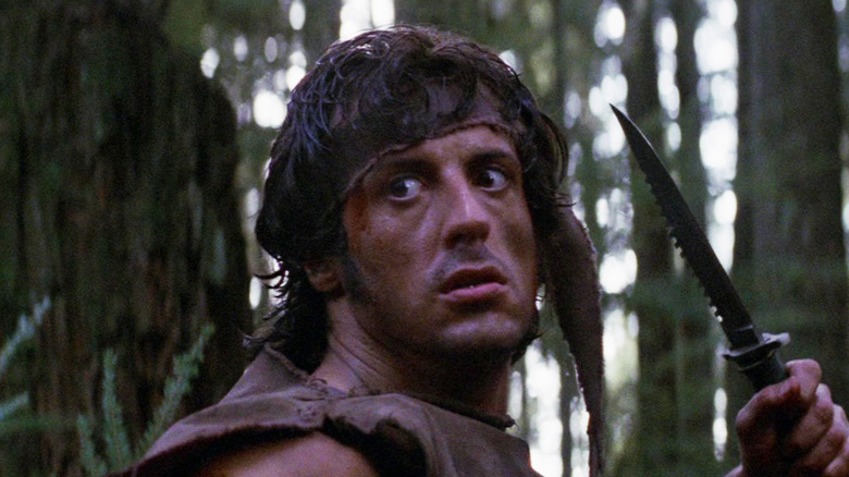 Sylvester Stallone's John J. Rambo holds a dagger while standing in a forest in First Blood