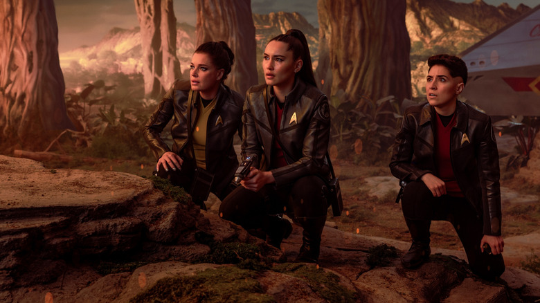 Rebecca Romijn as Una Chin-Riley crouching behind a rock in the wilderness next to Christina Chong as La'an Noonien-Singh and Melissa Navia as Erica Ortegas in Star Trek: Strange New Worlds
