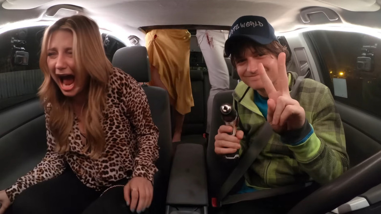 Joe Keery as Kurt Krunkle holding up two fingers in a peace sign and holding a drill while Mischa Barton as London screams in the seat next to him in Spree