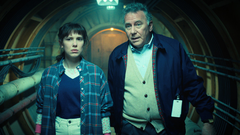 Millie Bobby Brown as Eleven walking down a hallway with Paul Reiser as Dr. Owens in Stranger Things