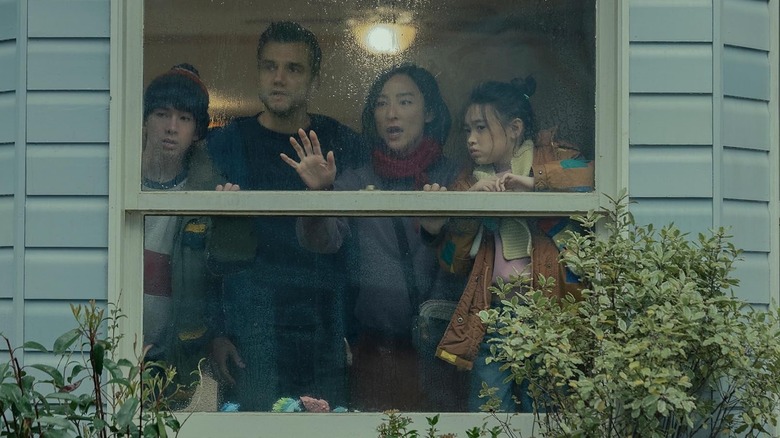 Greta Lee and Wagner Moura stand with children and look out the window of their house in The Last House