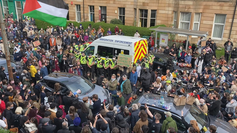 A crowd of protesters surround an immigration enforcement van on the street in Everybody to Kenmure Street