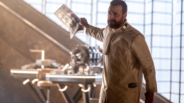 David Krumholtz as Zor-El holding a schematic and waving towards his work bench in Supergirl