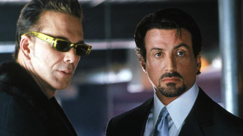 Mickey Rourke stands next to Sylvester Stallone's Jack Carter, who looks intensely at someone off screen in Get Carter
