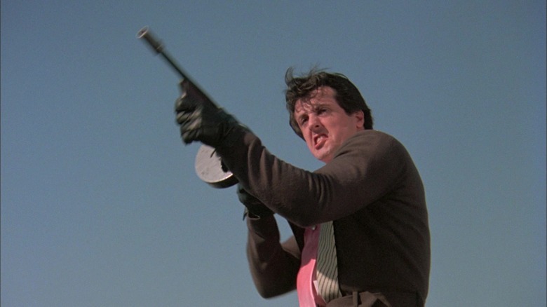 Sylvester Stallone's Joe Machine Gun Viterbo fires a rifle against a blue sky in Death Race 2000