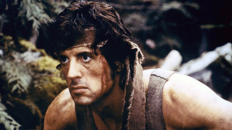 Sylvester Stallone as John Rambo in First Blood, with an intense look on his face as he stands in the forest