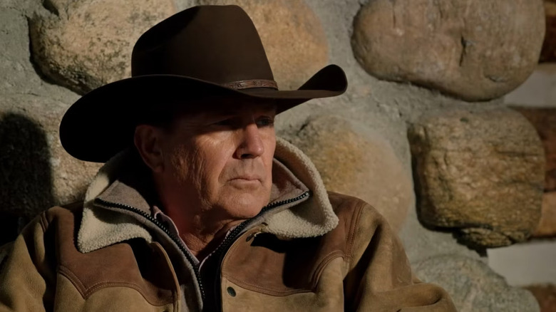 Kevin Costner as John Dutton sitting in front of a stone wall in his cowboy hat on Yellowstone
