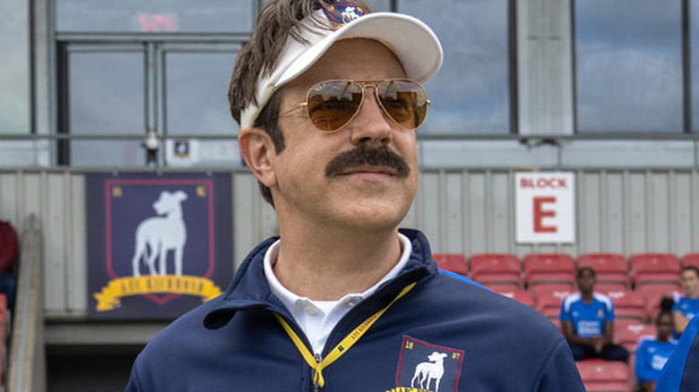Ted Lasso standing on the pitch with some players in bleachers behind him on Ted Lasso