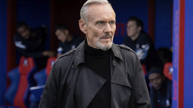 Rupert Mannion wearing a black jacket on the sidelines at a football match in Ted Lasso