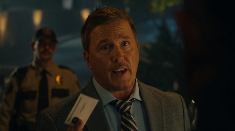 Lochlyn Munro as Larry Fitzgibbon handing Auggie his business card on Earth X on Peacemaker season 2