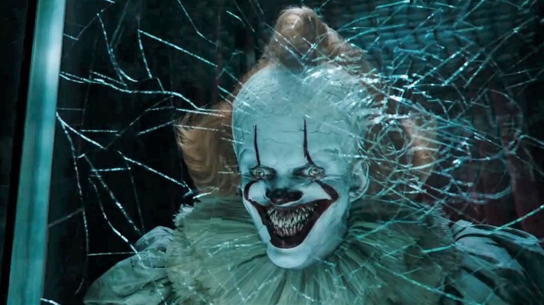Bill Skarsgard as Pennywise smiling behind a cracked window in It: Chapter Two