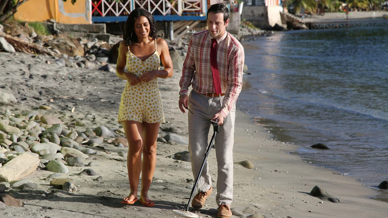 Neville Parker (Ralf Little) walks with a metal detector on the beach, accompanied by Florence Cassell (Joséphine Jobert) in Death in Paradise