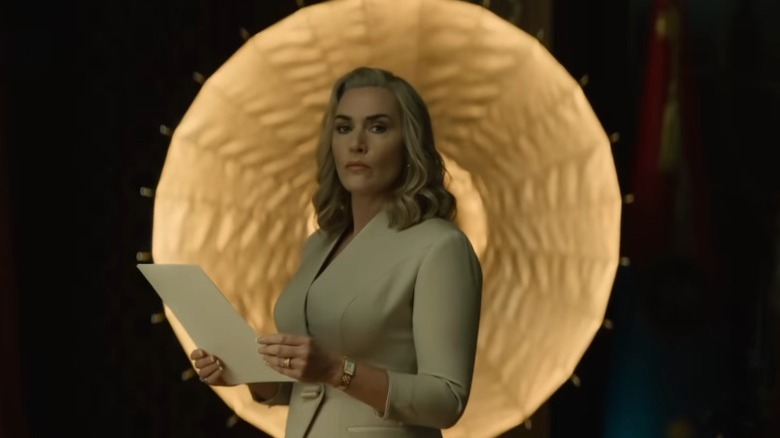 Kate Winslet as Elena Vernham holding papers and standing in front of a round decorative light fixture on The Regime