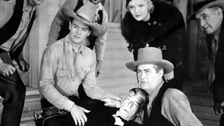 John Wayne as U.S. Marshal John Travers in The Star Packer, kneeling next to a fallen older man with other citizens, all looking out to something out of frame.