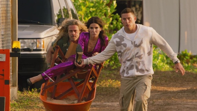 Brad Pitt as Jack Trainer pushes a wheel barrel carrying Sandra Bullock as Loretta Sage while Channing Tatum as Alan holds her foot in The Lost City