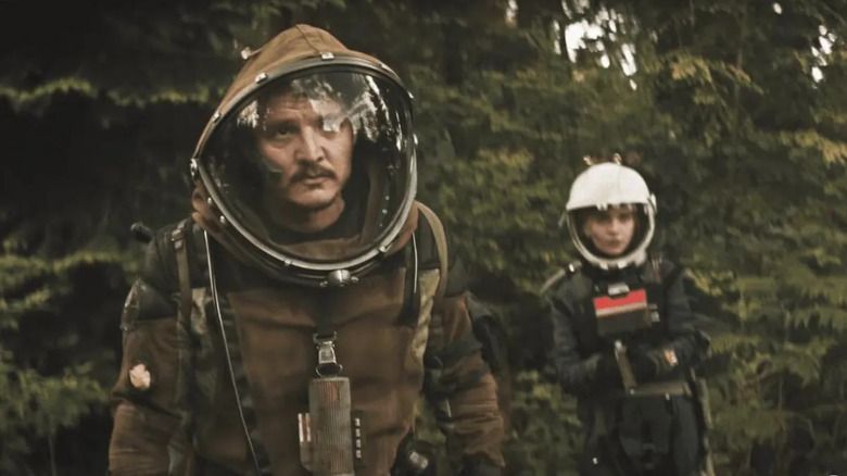 Ezra (Pedro Pascal) in a spacesuit stands with Cee (Sophie Thatcher) holds a gun behind him in Prospect