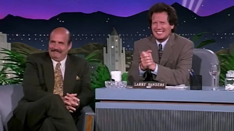 Larry Sanders (Garry Shandling) smiles behind his desk next to a seat Hank Kingsley (Jeffrey Tambor) in The Larry Sanders Show