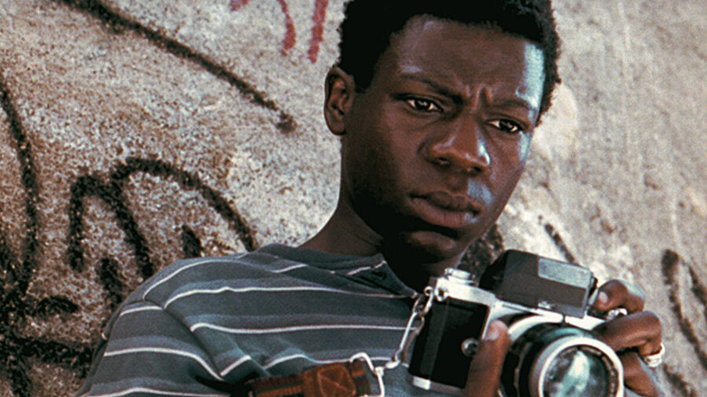Rocket (Alexandre Rodrigues) contemplatively holds his camera while leaning on a wall with graffiti in City of God