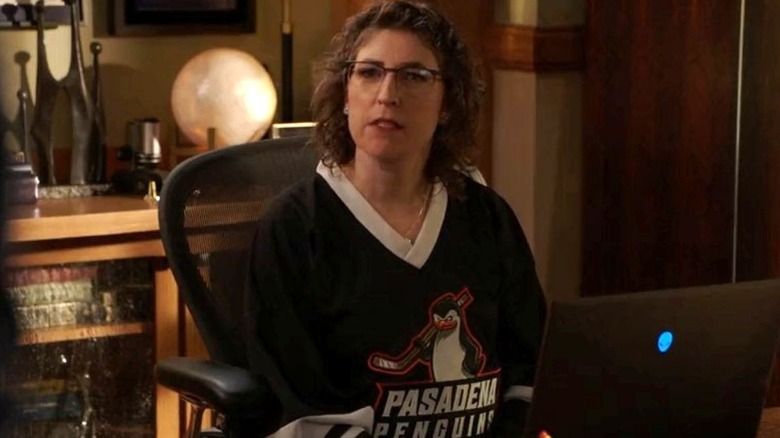 Mayim Bialik as Amy Farrah Fowler wears a hockey jersey while sitting at a desk on Young Sheldon