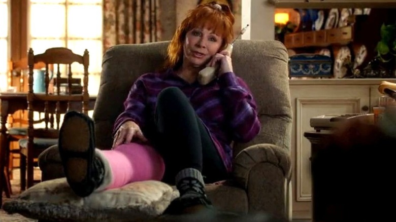 Reba McEntire as June sits in a chair with her leg in a cast while talking on the phone on Young Sheldon