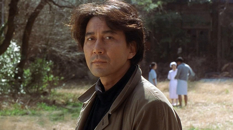 Kenichi Takabe (Kōji Yakusho) stands outside with hospital staff in the background in Cure