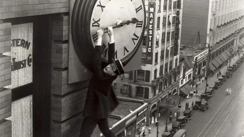 Harold Lloyd in glasess, suit, and hat hanging from large clock affixed to skyscraper in Safety Last!